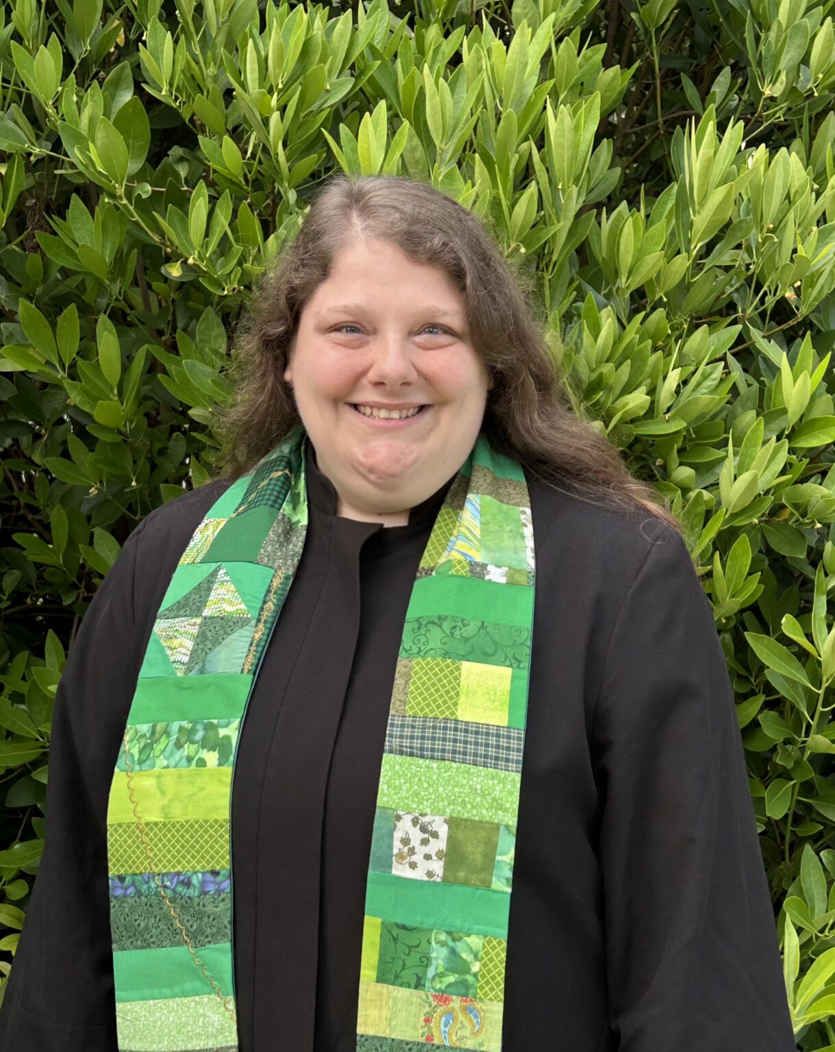 Rev. Whitney Hair in a black robe and green patterned stole. She is standing in front of a tall green bush.